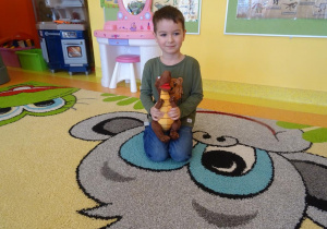 Chłopiec siedzi na dywanie, trzyma w ręku maskotkę dinozaura.